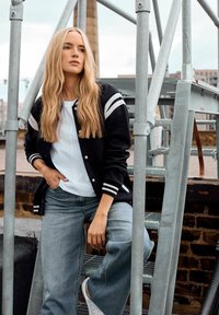 En blond kvinde iført en sort varsity-jakke, en hvid t-shirt og blå jeans sidder på udendørs metaltrapper med et urbant baggrund.