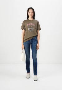 Oliven grønn grafisk t-skjorte med løveprint, parret med blå denimjeans og grå slip-on sko. Holder en liten hvit håndveske.