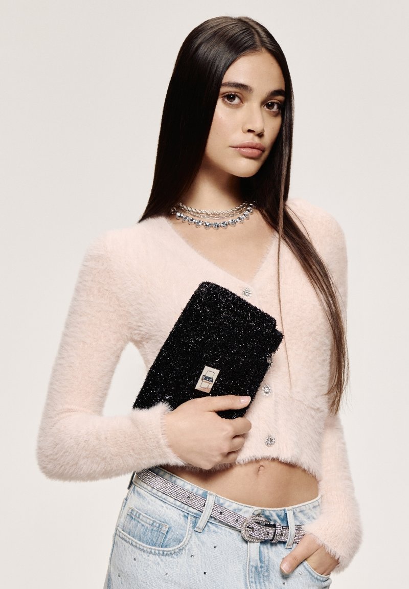 Cardigan corto rosa fluffy con maniche lunghe, bottoni decorativi, abbinato a jeans azzurri e una pochette nera scintillante.