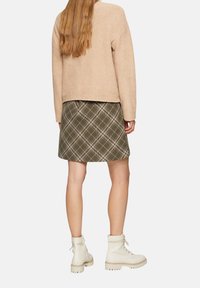 Beige strukturierter Pullover, gemusterter grüner Karorock, weiße Knöchelstiefel mit Schnürung und klobigen Sohlen, von hinten betrachtet.