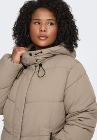 Beige puffjacka med hög krage, justerbara dragsnören och sidofickor. Har en vadderad design och texturerat tyg.