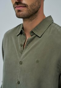 Olive zelena srajca iz teksturiranega blaga, z ovratnikom na gumb, izvezenim logotipom na prsih ter vidnimi gumbi in šivnimi detajli.