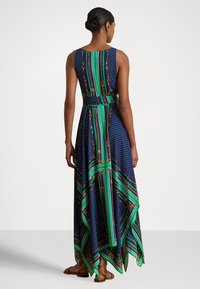 Γυναίκα που στέκεται με την πλάτη ορατή, φορώντας ένα αμάνικο μπλε σκούρο φόρεμα με πράσινα και καφέ ριγέ και πουά σχέδια και με ασύμμετρη τελείωμα σαν μαντήλι.