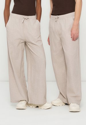 Dwie osoby w beżowych spodniach o szerokich nogawkach ze sznurkami i białych trampkach, stojące obok siebie na jednolitym tle.