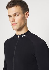 Maillot de cyclisme noir à manches longues en tissu extensible. Il présente un col haut, une fermeture éclair sur le devant et un logo au niveau du col.