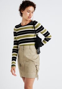 Csíkos, bordázott, hosszú ujjú felső olíva zöld, fekete és krémszínben, bézs cargo szoknyával párosítva, amelyen hasznos zsebek és zsinóros szegély található.