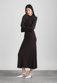 Vestido ajustado de manga larga en marrón oscuro, con cuello alto, abertura lateral y textura suave. Llevado con tacones blancos de punta.