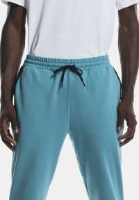 Personne à la peau foncée portant un pantalon de jogging bleu clair avec des cordons de serrage noirs et des rayures latérales, associé à un t-shirt blanc uni, les mains détendues.
