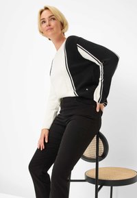 Schwarz-weiß gestrickter Pullover mit diagonalen Streifen, ausgestattet mit gerippten Bündchen und einem strukturierten Muster, kombiniert mit schwarzen Hosen und einem geflochtenen Stuhl.