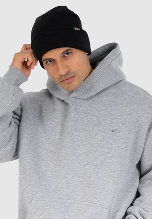 Grå hættetrøje med en kængurulomme og et lille mærketik. Sort ribbet beanie. Model som ser lidt til siden med en hånd på hovedet.