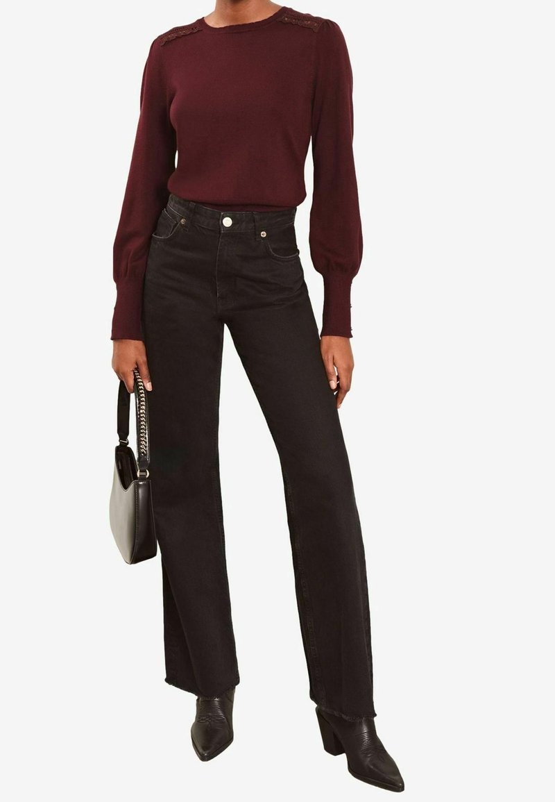 Femme portant un haut bordeaux à manches longues, un jean noir taille haute à jambes larges, des bottines noires à talons, tenant un sac à main noir.