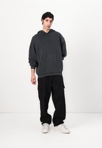 Tamsiai pilka hoodie, su laisvu siluetu ir kengūros kišene, derinama su juodomis kargo kelnėmis, turinčiomis šonines kišenes, ir baltais sportiniais batais.