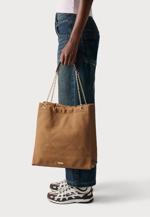 Tan quilted tote bag with a cinched top, chain-link handles, and a smooth texture, paired with denim jeans and sneakers.