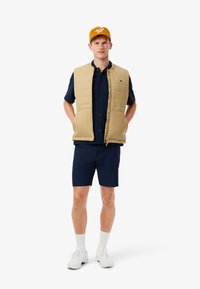 Gilet imbottito beige sopra una maglietta blu navy e pantaloni corti. Il modello indossa scarpe bianche e un cappellino giallo con un logo. Il gilet presenta una cerniera frontale e tasche.