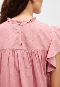 Ružičasta pamučna bluzica s ruffles izrezom, kratkim rukavima i teksturiranim uzorkom točkica. Sadrži otvor u obliku ključa s dugmetom na leđima.