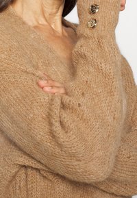 Cardigan de mohair beige con diseño de cuello en V, puños acanalados con dos acentos de botones dorados, y un acabado esponjoso y texturizado.