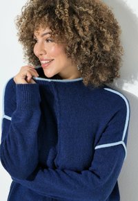 Frau mit lockigem Haar, die lächelt und einen blauen Pullover mit hellblauen Paspeln trägt, während sie den Kragen nahe ihres Gesichts hält, vor einem hellen Hintergrund.