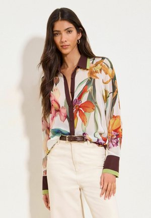 Kobieta ubrana w przezroczystą kwiecistą bluzkę z ciemnym obszyciem i kremowe spodnie z wysokim stanem, stoi na tle jednolitego jasnego tła.