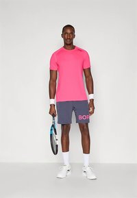 Homem em pé com uma camisa rosa, calções cinzentos com texto rosa, pulseiras e sapatos brancos, segurando uma raquete de ténis contra um fundo simples.