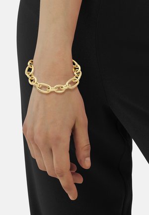 Bracelet en chaîne dorée présentant des formes ovales et rondes entrelacées, finition polie, reposant sur une main. Fond en tissu noir lisse.
