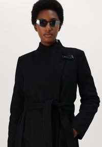 Cappotto nero con colletto alto, tasche laterali e cintura annodata. Presenta un dettaglio di chiusura unico sul davanti. Il modello indossa occhiali da sole neri angolari.