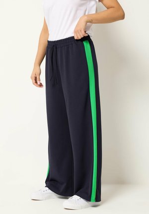 Personne portant un pantalon noir ample avec des bandes latérales vertes, des baskets blanches et une chemise blanche à manches courtes, debout devant un fond uni.