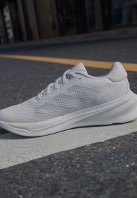Zapatilla deportiva blanca con parte superior de malla, suela texturizada y tres rayas laterales sutiles. Diseño redondeado con un talón acolchado para mayor soporte.