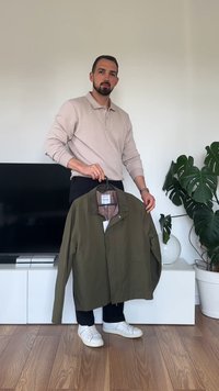 Olivno zeleni plašč z ovratnikom, sprednjimi pritisnili in karo podlogo. Model drži oblačilo na obešalniku, oblečen v svetel polo.