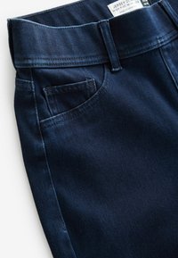 Jean en denim bleu foncé taille haute avec jambes larges, poche avant visible, passants de ceinture et étiquette partiellement visible à l'intérieur de la ceinture.
