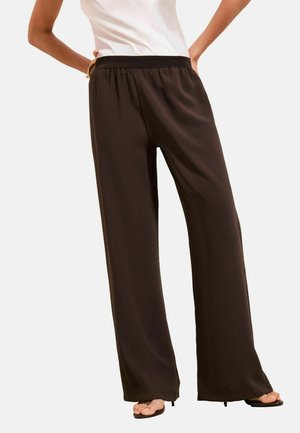 Femeie purtând pantaloni largi maro închis cu talie elastică și sandale negre cu degetele descoperite, stând cu mâinile pe șolduri.