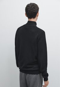 Zwarte pullover trui met een hoge kraag, geribbelde textuur langs de schouders en mouwen, en een ontspannen pasvorm. Glad stof en strakke vormgeving.