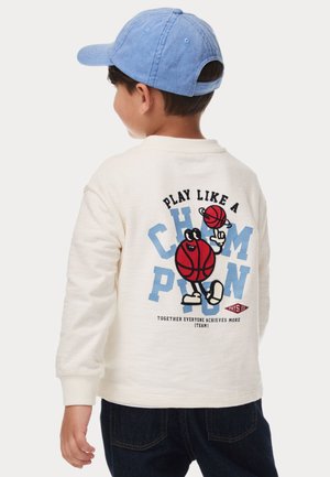 White textured pullover featuring a colorful cartoon graphic of a basketball player with text. Blue cap contrasts with dark denim jeans.