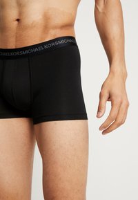 Crne pamučne bokserice s širokim crnim pojasom na kojem je u srebrnom slovu ispisano "MICHAEL KORS". Tkanina pokazuje laganu rastezljivost i glatku teksturu.
