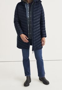 Granatowa pikowana kurtka puchowa z kapturem, z zamkiem z przodu, wydłużonym fasonem i bocznymi kieszeniami, noszona na koszulę dżinsową i dżinsy.