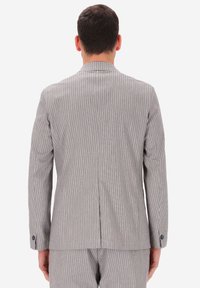Uomo di spalle con indosso un giacca di completo grigio chiaro a righe e pantaloni abbinati, su uno sfondo bianco.
