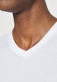 Close-up van de nek en bovenste borst van een persoon die een wit T-shirt met V-hals draagt tegen een effen achtergrond.