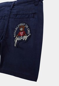 Marineblaue Shorts mit einem gestickten Bärenlogo, versehen mit dem Text "Gegründet 1981" und "Guess", detaillierter Nahtführung und zwei Gesäßtaschen.