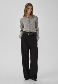 Szürke kötött kardigán gombos elől, laza fekete nadrággal és széles fekete övvel, amelyen fémből készült csat látható.