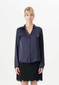 Woman wearing a long-sleeve navy satin blouse with buttons and a black skirt with a scalloped silver sequin hem.