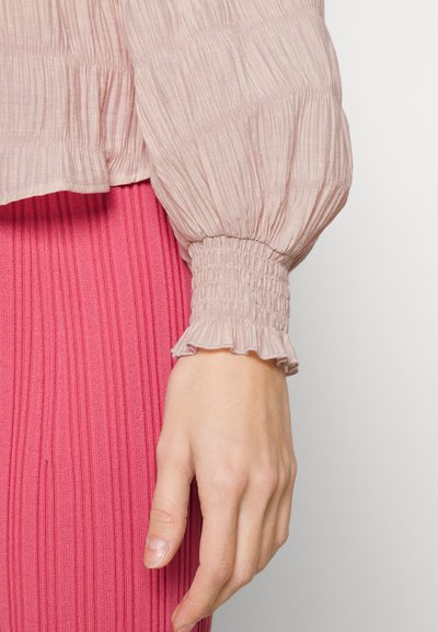 Χέρι γυναίκας resting στο πλάι, φορώντας ροζ μπλουζάκι με υφή και gathered cuff, και φωτεινή ροζ φούστα με ρίγες.