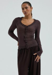 Femme portant un haut moulant à manches longues marron foncé avec des bords volantés et un pantalon ample marron foncé, debout devant un fond clair uni.