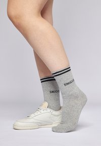 Graue Socken mit schwarzen Streifen am oberen Rand, mit dem Text "SNOCKS". Getragen zu weißen Sneakers vor einem neutralen Hintergrund. Glatte Textur.