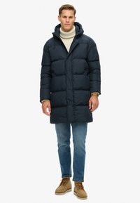 Abrigo acolchado azul marino con capucha, que presenta un diseño acolchado, cierre frontal y bolsillos laterales, combinado con jeans de color azul claro y botas marrones.