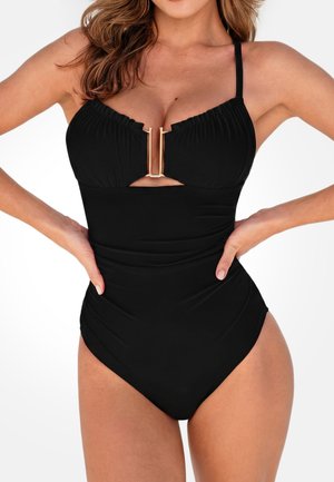 Vrouw met een zwarte eendelige badpak met een gouden U-vormige sluiting aan de voorkant en een halternek, handen op de heupen tegen een witte achtergrond.