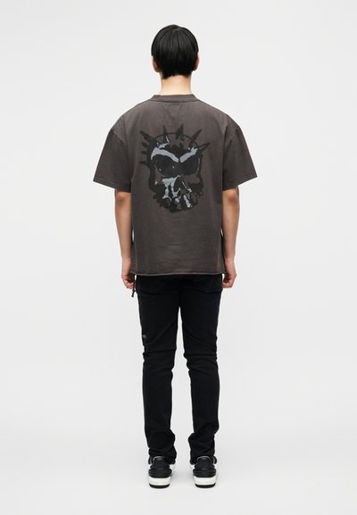 Γκρι φαρδιά μπλούζα με μεγάλο μαύρο γραφικό κρανίου στην πλάτη, σε συνδυασμό με μαύρο στενό παντελόνι και μαύρα αθλητικά παπούτσια.