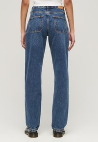 Pantalones vaqueros de mezclilla azul con corte recto, bolsillos traseros visibles y una etiqueta de cuero en la cintura. Tejido texturizado de peso medio.