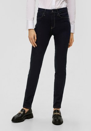 Donkerblauwe skinny jeans van denim met een hoge taille, enkele knoopsluiting en gouden stiksels. Gecombineerd met zwarte loafers met gouden accenten.