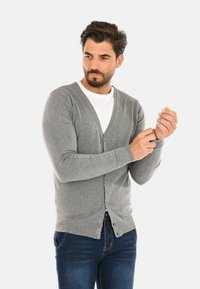 Cardigan grigio lavorato a maglia con scollo a V, polsini e orlo a coste, con bottoni sul davanti. Indossato sopra una maglietta bianca e jeans blu.