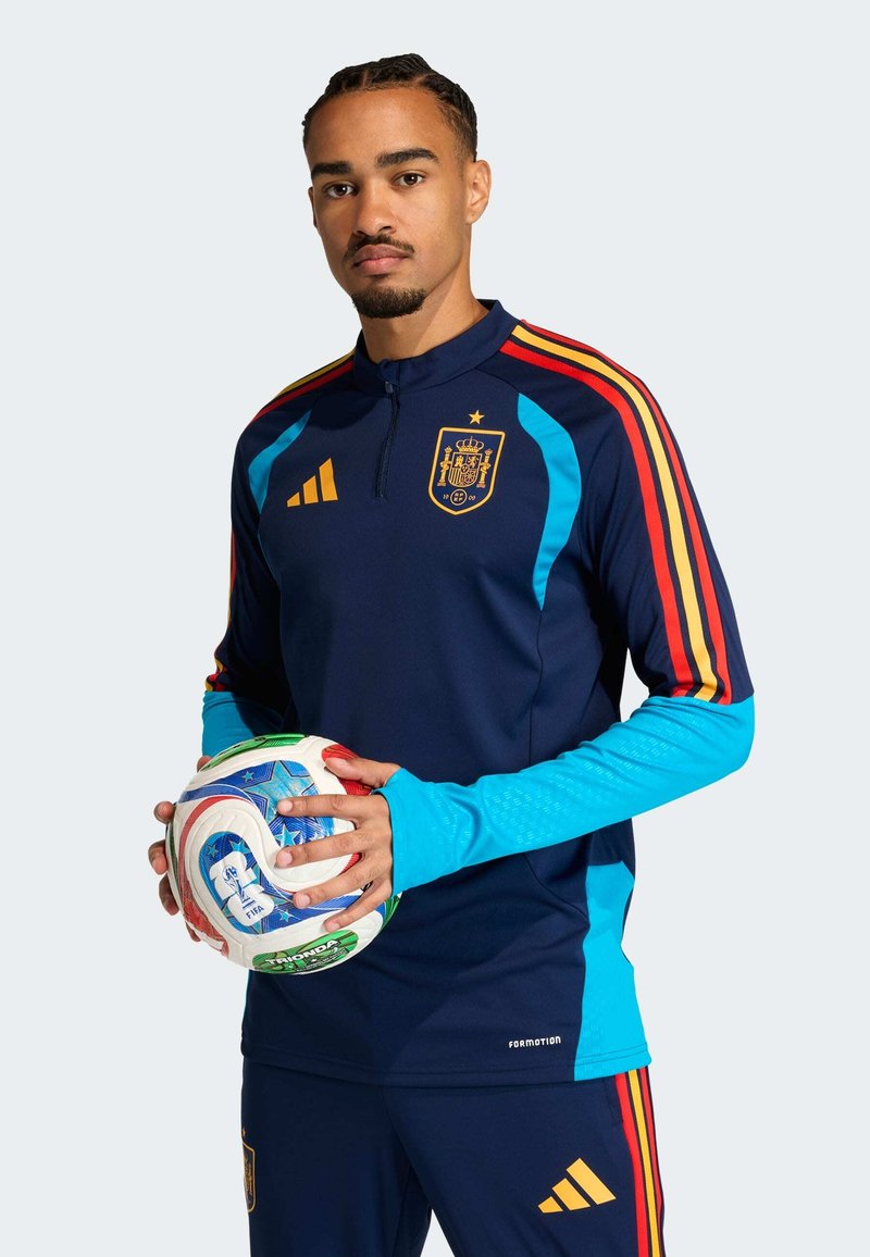 Camisola de treino da seleção nacional de Espanha em azul-marinho com detalhes em vermelho e amarelo, com gola de meio fecho e mangas azul-claro. Segurando uma bola de futebol multicolorida.