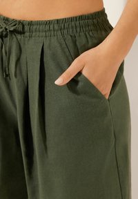 Pantalones de tela verde, ligeros, con cintura elástica, bolsillos laterales y pliegues, que exhiben un diseño relajado y una textura suave.
